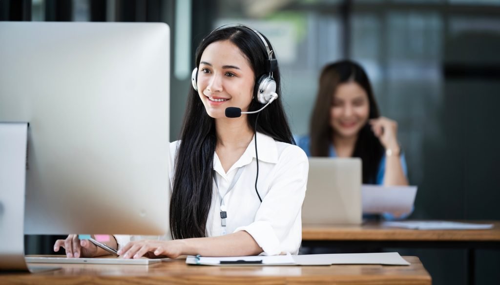 Smiling Asian female operator speaking with client via headset while working in call center